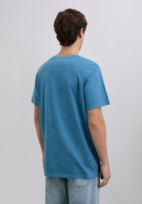 Ung mand med kort brunt hår iført en enkel blå T-shirt med korte ærmer og lyseblå jeans, står vendt væk mod en ensfarvet baggrund.