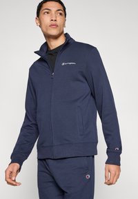 Μπλε σκούρο φούτερ με φερμουάρ και ψηλό γιακά, με μικρό λευκό λογότυπο στο στήθος και δύο πλαϊνές τσέπες. Μαλακό, υφηρό ύφασμα.