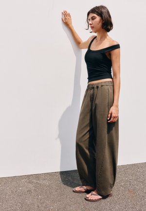 Jeune femme aux cheveux courts bruns portant un haut noir sans manches, un pantalon large olive et des tongs, debout en touchant un mur blanc.