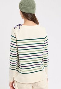 Femme portant un haut à manches longues crème avec des rayures horizontales vertes, violettes et teal, un pantalon beige et un bonnet en tricot vert olive, de dos.