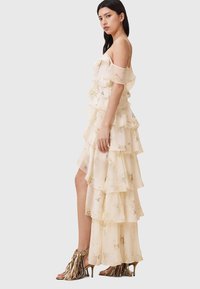 Lagdelte, rynkede kjole i lys creme med blomstermønster. Spaghetti-stropper og asymmetrisk kant. Sølvmetallic hælede sandaler.