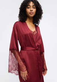 Γυναίκα που φοράει ρόμπα από βουργουνδί σατέν με δαντέλα στα μανίκια και αντίστοιχη καμιζόλα με δαντέλα από κάτω, στέκεται μπροστά σε ένα απλό φόντο.