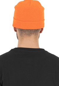 Oransje ribbestrikket lue med brettet oppkant, båret på hodet til en person. Svart skjorte synlig i forgrunnen.
