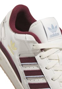 Witte leren sneaker met bordeauxrode accenten, voorzien van drie bordeauxrode strepen en een blauwe adidas-logo op de tong. Gekorrelde rubberen zool.