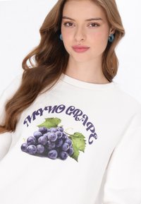 Hanorac alb cu un grafic cu struguri mov cu frunze verzi și textul „NONO GRAPE” în mov închis, cu guler rotund, material moale.