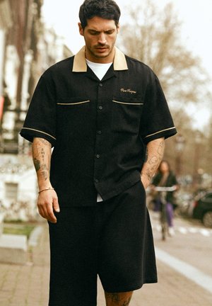 Bărbat cu tatuaje purtând o cămașă cu guler negru și maro deschis și pantaloni scurți negri, mergând pe stradă în aer liber, cu un ciclist neclar în fundal.