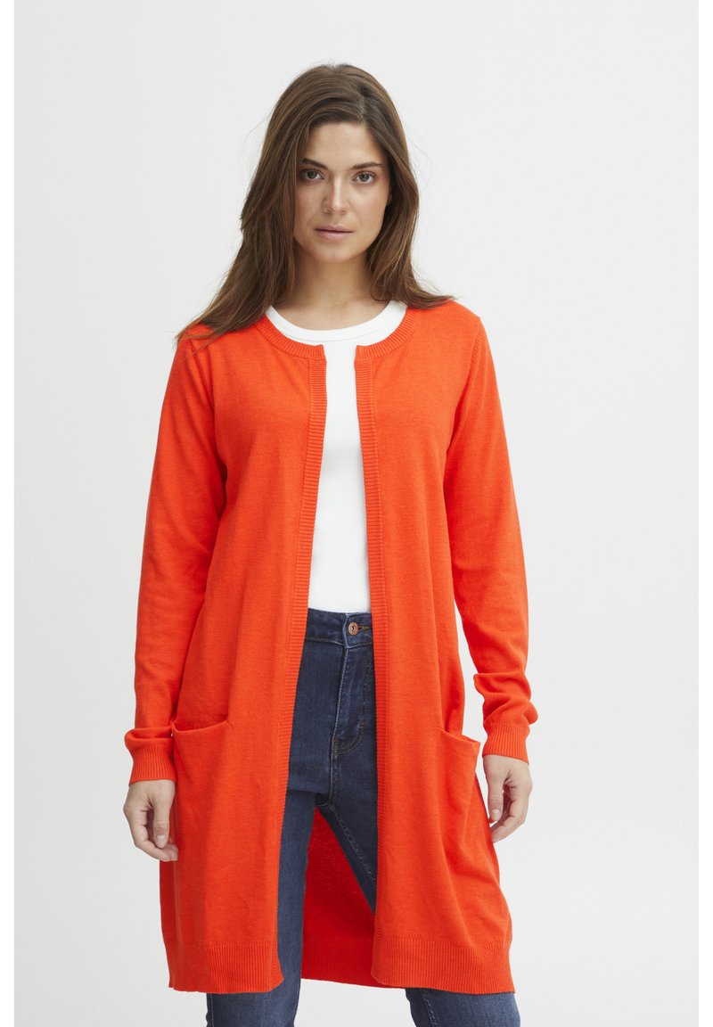 Long gilet orange avec des finitions côtelées et deux poches avant, porté par-dessus un haut blanc, assorti avec un jean bleu foncé. Texture tricot lisse.