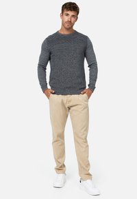 Grauer strukturierten Strickpullover mit rundem Ausschnitt, kombiniert mit hellbeigen Chinos und weißen Sneakern, Hände in den Taschen, neutraler Hintergrund.
