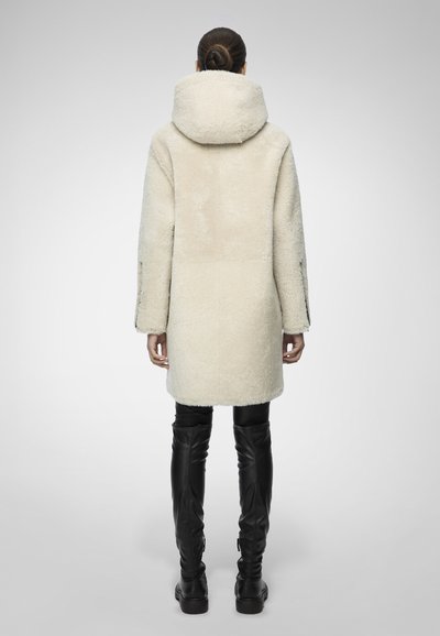 Donna con capelli raccolti in uno chignon, che indossa un cappotto di shearling color crema lungo fino al ginocchio e stivali in pelle nera alti fino alla coscia, in piedi di spalle su uno sfondo grigio.