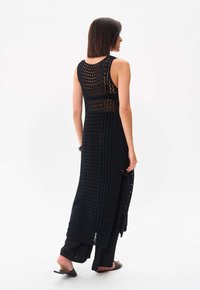 Schwarze Häkelstrick-Weste mit langem Design und ärmelloser Schnittführung, ausgestattet mit einem offenen Rücken und seitlichen Schlitzen. Kombiniert mit schwarzen Hosen und Sandalen.