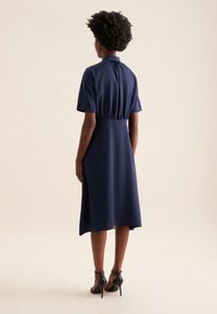 Abito blu navy con maniche corte, schiena arricciata e silhouette a trapezio lunghezza ginocchio. Indossato con scarpe con tacco alto nere.