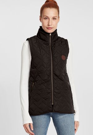Gilet matelassé noir avec col montant, fermeture éclair à l'avant et poches latérales. Présente un patch logo marron sur la poitrine gauche. Porté sur une chemise blanche.