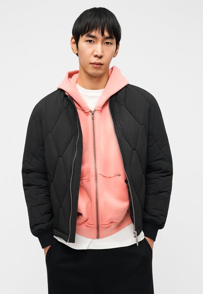 Μαύρο παλτό bomber με καπιτονέ ύφασμα πάνω από ροζ φούτερ με φερμουάρ, με λευκό μπλουζάκι λαιμόκοψης και μαύρο παντελόνι, εμφανιζόμενο από μπροστά.