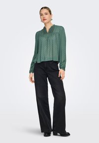 Γυναίκα όρθια, φοράει διάφανη πράσινη μπλούζα με μακριά μανίκια και δαντελένιες λεπτομέρειες, μαύρο τζιν με φαρδιά πόδια και μαύρα παπούτσια.