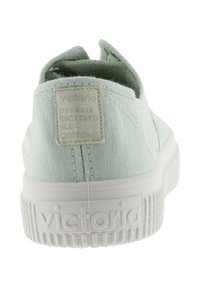 Zapatilla de lona verde claro sin cordones con suela de goma blanca, patrón texturizado en la suela y una etiqueta que dice "Victoria" en la parte trasera.