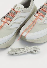 Light gray sneakers with white accents, textured material, and orange patterned laces. Includes additional gray laces beside the shoes.