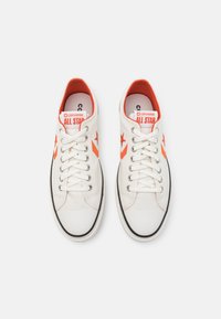 Zapatillas de lona blancas con detalles en naranja, puntera de goma y ribete negro. Presentan cordones tradicionales y un logo de estrella en el lateral.