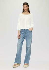 Weißer gestrickter Pullover mit U-Ausschnitt und strukturiertem Muster, kombiniert mit hochgeschnittenen hellblauen Jeans und Plateau-Sandalen.