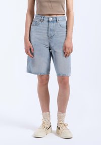 Pantalones cortos de mezclilla azul claro con cintura alta y corte recto, que cuentan con cinco bolsillos y detalles en botones metálicos. Combinados con zapatillas beige y calcetines.