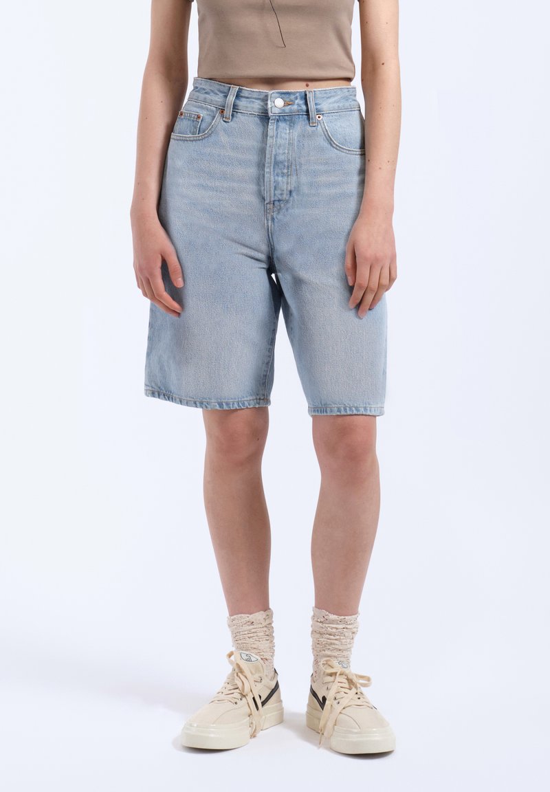 Pantalones cortos de mezclilla azul claro con cintura alta y corte recto, que cuentan con cinco bolsillos y detalles en botones metálicos. Combinados con zapatillas beige y calcetines.