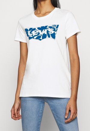 Vrouw die een wit Levi's t-shirt draagt met een blauw patroonlogo, gecombineerd met lichtblauwe spijkerbroek, staand tegen een effen achtergrond.