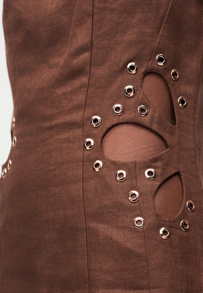 Καφέ ύφασμα με διακοσμητικές οπές περιβαλλόμενες από μεταλλικά οδηγούς που σχηματίζουν ένα αφηρημένο μοτίβο σε ένα ένδυμα.