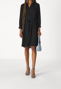 Svart knapprad klänning med långa ärmar, insvängd midja och rak passform. Kombineras med blå klacksandaler och en liten blå väska.