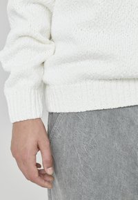 Pull en maille blanche avec un tissu texturé et des poignets côtelés, associé à un pantalon en denim gris, illustrant une tenue décontractée.