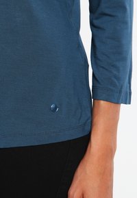 Tyrkysová košile s dlouhým rukávem a kulatým výstřihem. Součástí je malý modrý logo detail na lemování a hladký, mírně pružný materiál.