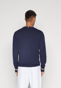 Maglione blu navy con maniche lunghe, polsini a coste con dettagli rossi e bianchi, e scollatura a girocollo, mostrato da dietro.