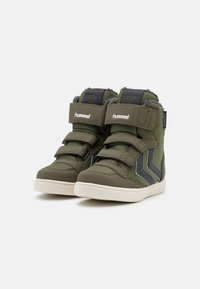 Un par de zapatillas altas para niños en color verde oliva con suelas blancas, correas de velcro y detalles en chevron gris en los costados.