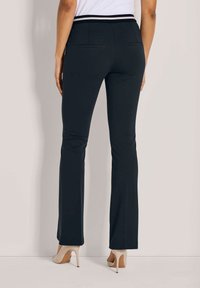 Vrouw in zwarte aansluitende rechte broek en beige hoge hakken, staand tegen een egale lichte achtergrond.
