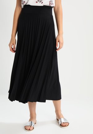 Mujer vistiendo una falda midi plisada negra, blusa floral metida por dentro y sandalias plateadas abiertas en los dedos, de pie contra un fondo blanco liso.