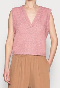Haut en tricot sans manches de couleur rose avec un col en V, présentant une finition douce et texturée. Porté par-dessus un pantalon large beige avec des poches.