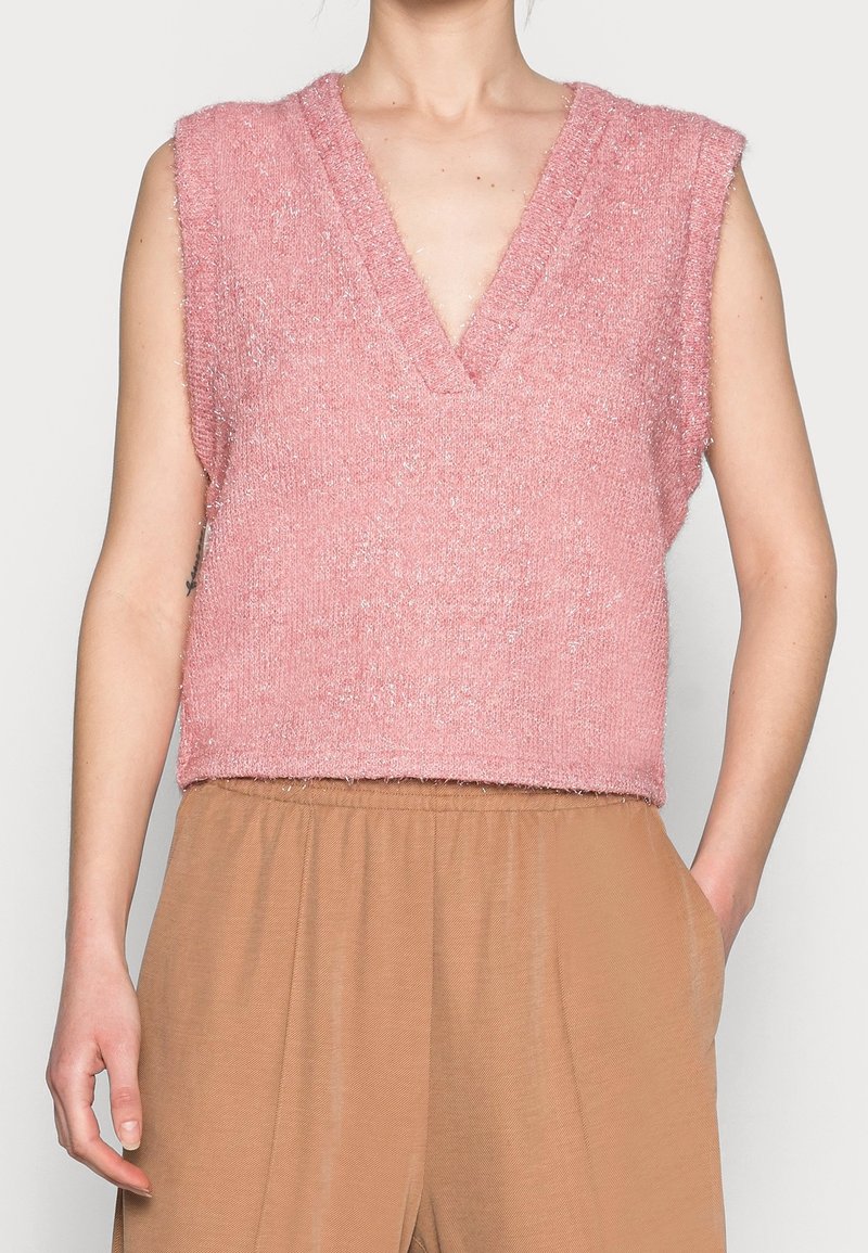 Haut en tricot sans manches de couleur rose avec un col en V, présentant une finition douce et texturée. Porté par-dessus un pantalon large beige avec des poches.
