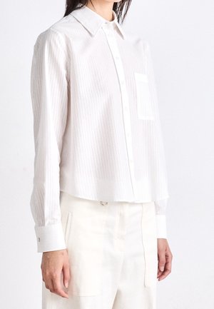 Femme portant une chemise blanche à manches longues avec poche poitrine et un pantalon taille haute écru, se tenant devant un fond blanc.