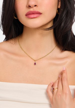 Goldene Halskette mit einem tropfenförmigen roten Edelsteinanhänger und floralem Akzent. Glatte Hautfarbe, an der Hand hellrosa Nagellack.