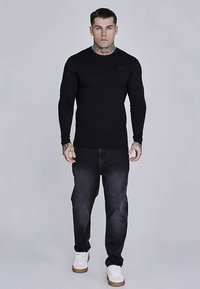 Crna uska košulja s dugim rukavima i rebrastom teksturom, uparena s tamnim trapericama i bijelim tenisicama. Jednostavan dizajn s malim logotipom na prsima.