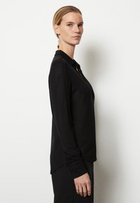 Camisa negra de manga larga con botones y cuello, hecha de un material suave, con corte recto y una ligera cola en la parte posterior.