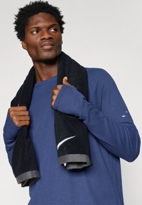 Μπλε πουκάμισο με μακριά μανίκια και ριπ ανάγλυφη υφή, συνδυασμένο με μια μαύρη πετσέτα που διαθέτει λευκό λογότυπο. Χέρια κρατούν την πετσέτα, δείχνοντας σφιχτή εφαρμογή.