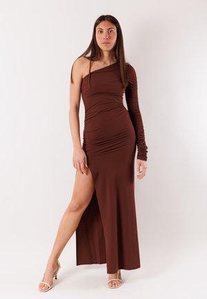 Donna con abito marrone monospalla a manica lunga con spacco sulla coscia e tacchi beige aperti in piedi su sfondo bianco.