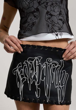 Personne portant une mini-jupe noire avec des lettres gothiques blanches et un crop top en mesh noir sur un t-shirt blanc, les mains ajustant la ceinture de la jupe.