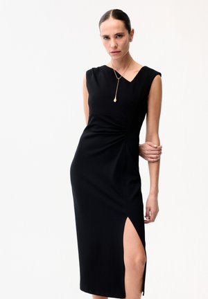 Kobieta w bezrękawnikowej czarnej sukience z rozcięciem do uda, nosząca wiszącą naszyjnik, stojąca na jednolitym białym tle.