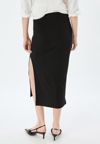 Jupe noire en jersey avec une fente haute sur le côté gauche, longueur genou, design ajusté. Portée avec des talons ouverts noirs.