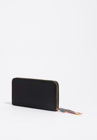 Cartera de cuero negro con textura suave, un zipper dorado y un adornado borlón con acentos rojos y grises. Forma rectangular.
