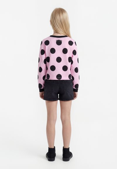 Dziecko z długimi blond włosami, ubrane w różową bluzkę z długim rękawem w duże czarne grochy, czarne dżinsowe spodenki i czarne buty, zwrócone tyłem.