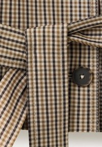 Gros plan d’un tissu à carreaux marron, beige et noir avec une ceinture nouée et un bouton foncé cousu sur le textile.