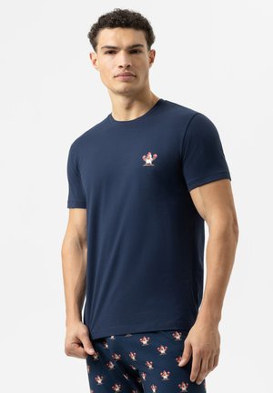 Mann in navyblauem T-Shirt und Hose mit kleinen Cartoon-Boxhahn-Drucken, der vor einem weißen Hintergrund steht.