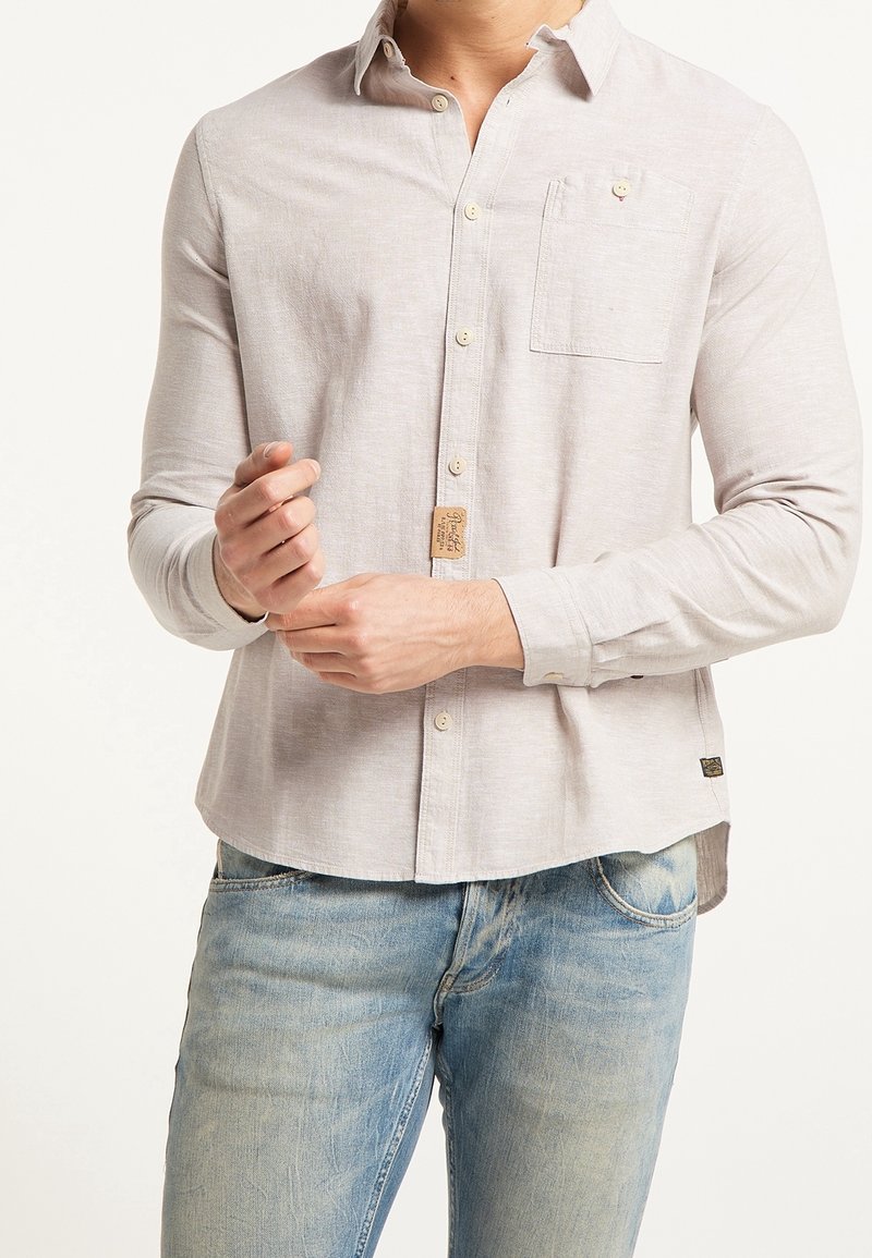 Homme portant une chemise beige clair à manches longues avec boutons et un jean bleu délavé, ajustant sa manche gauche avec sa main droite.