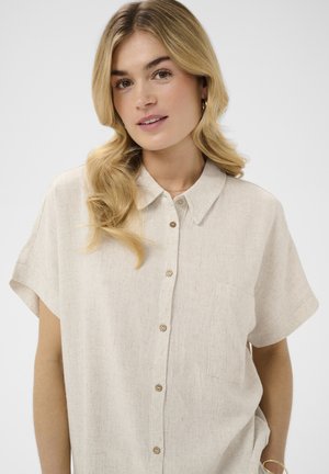  Heleblond naine, kellel on lahtised lokid, kannab beežikat lühikeste varrukatega nööpidega särki esitaskuga ning seisab ühevärvilise tausta ees.